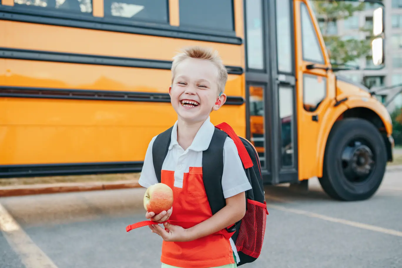 Jeune garçon portant des appareils auditifs et souriant devant un autobus scolaire