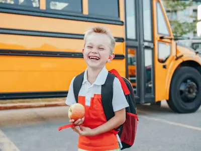 School aged boy wearing hearing aids and standing in front of a school bus smiling